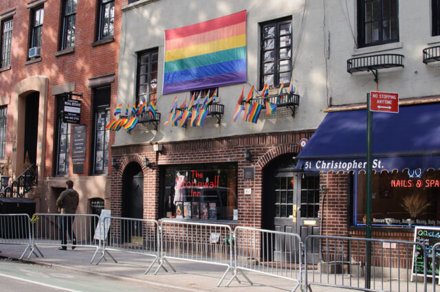 Front view of the Stonewall Inn in New York, adorned with a large rainbow flag and multiple smaller rainbow flags. The historic building is located at 51 Christopher Street and features a sign reading ‘The Stonewall Inn’ in its front window. Metal barricades are set up on the sidewalk, and a person is standing nearby. The scene reflects the significance of the location in LGBTQ+ history.