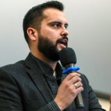 Dave Williams, chair of the Colorado GOP and candidate for Colorado Congressional District 5, speaking into a microphone. He has a beard, dark hair, and is wearing a dark suit jacket with a checkered shirt. The background is plain and neutral, focusing attention on Williams as he speaks.