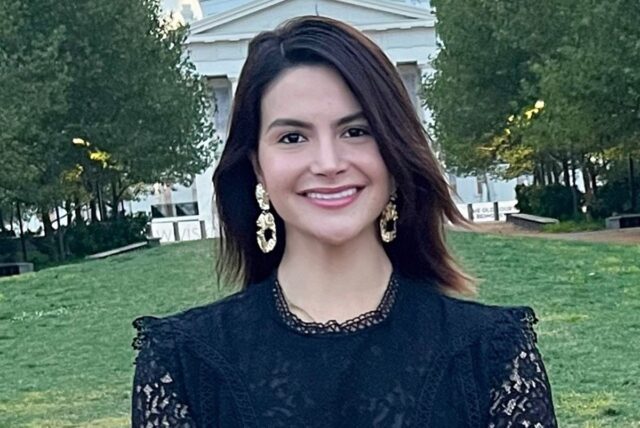 Portrait of Valentina Gomez, a candidate for Missouri Secretary of State, smiling and wearing a black lace top with gold earrings, with a park and building in the background.