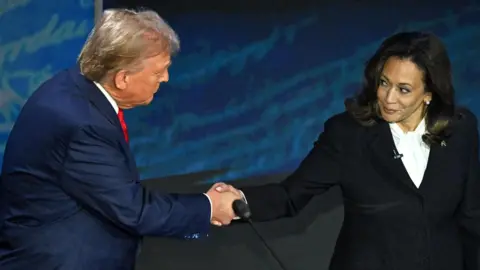 Donald Trump and Kamala Harris shake hands on stage during a political event. Trump, wearing a navy blue suit and red tie, reaches across to shake hands with Harris, who is dressed in a black suit and white blouse. Harris looks at Trump with a composed expression while Trump leans slightly forward, meeting her gaze. The background features a blue backdrop with indistinct writing or design elements.